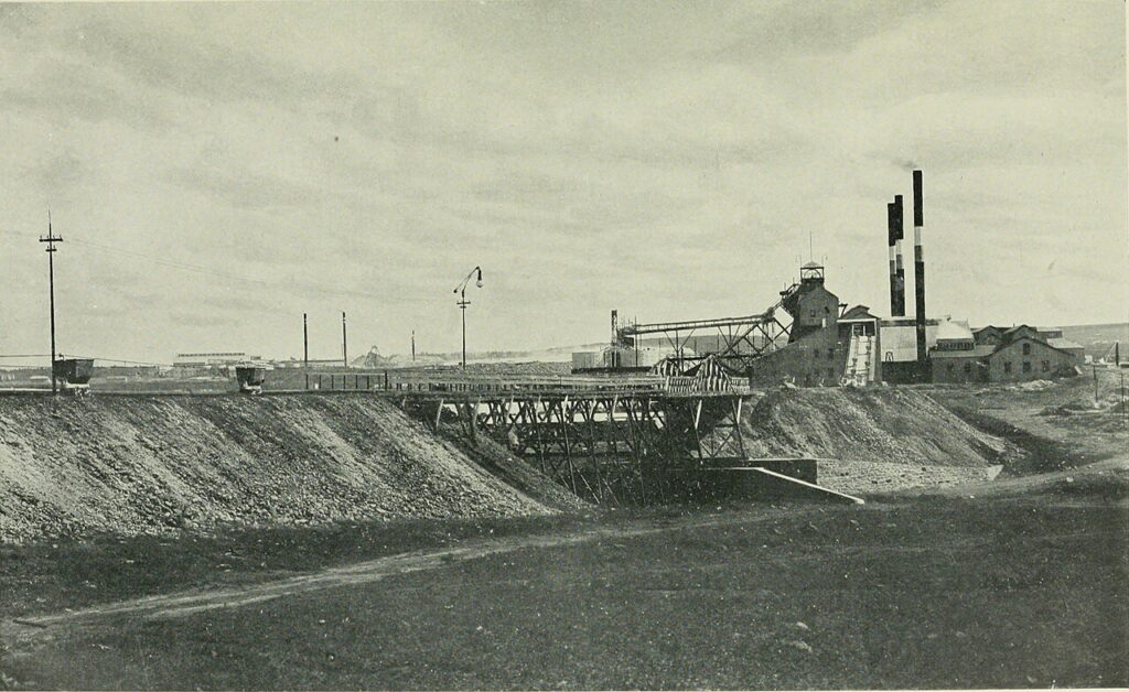 A disused mining site with a narrow shaft entrance set into rocky, sandy terrain under a pale sky, with weathered wooden supports visible.