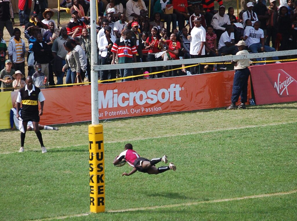 An amateur rugby match in progress on a grass pitch in an African setting, with players in contrasting kits contesting open play and a small