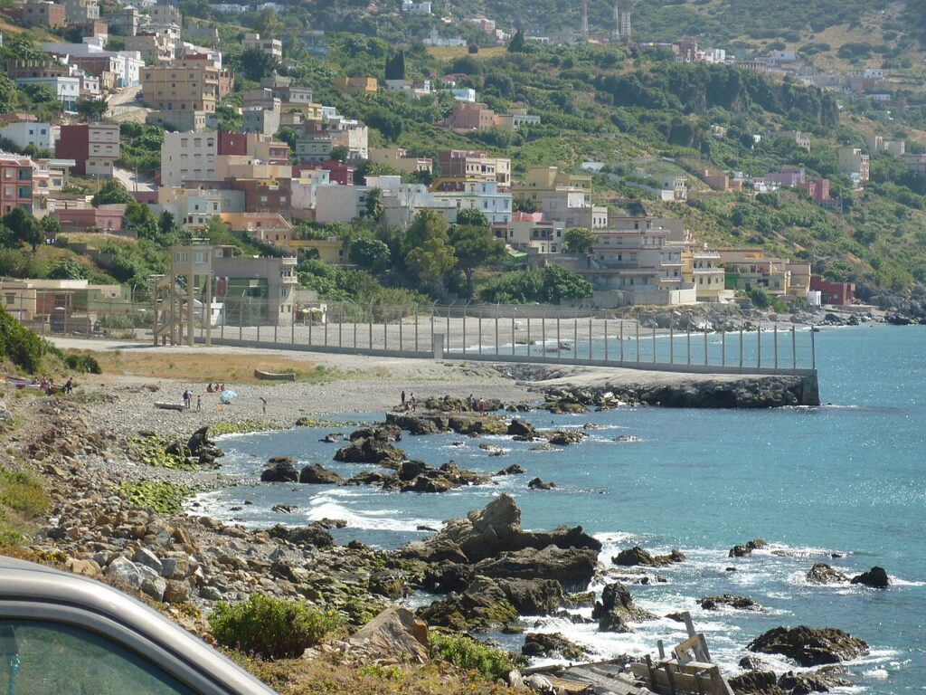 A fortified double border fence with a patrol road running alongside, in a coastal Mediterranean setting with security personnel visible at