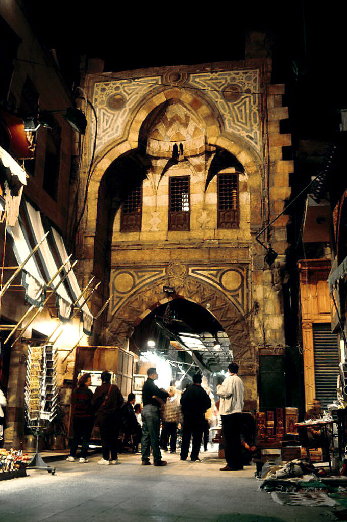 A lively bazaar alley lined with souvenir stalls as a man in casual clothing interacts warmly with shopkeepers and visitors.