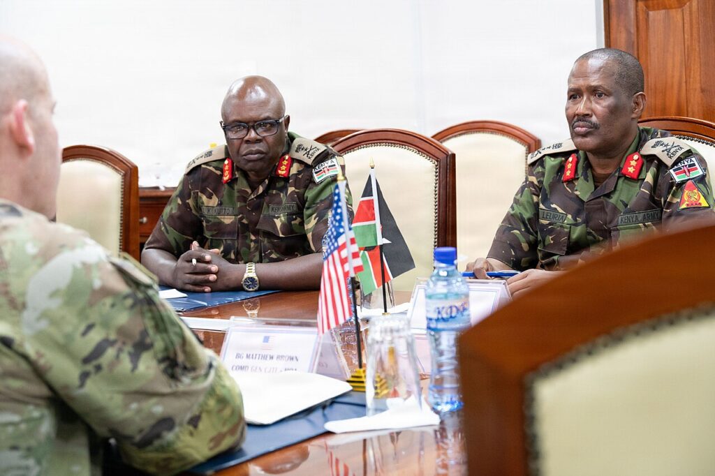 A formal diplomatic meeting between two defence delegations seated across a conference table with national flags displayed in the background