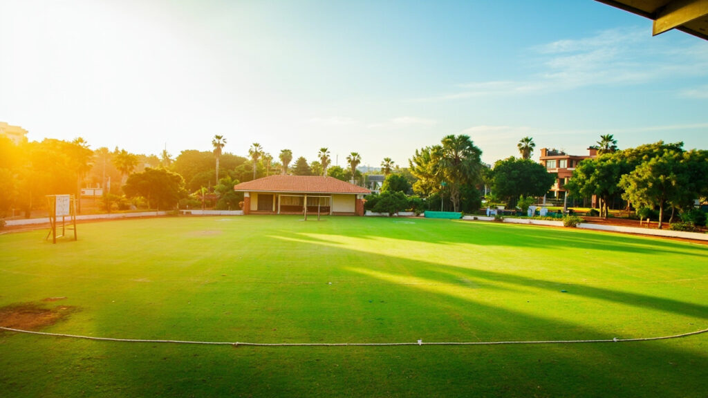 A cricket oval at a university sports ground with a central pitch, pavilion-style clubhouse and acacia trees visible in the background under