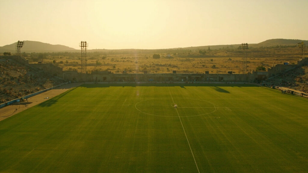 A modest football stadium in the Singida region of Tanzania, with a green grass pitch and simple concrete stands under open sky.