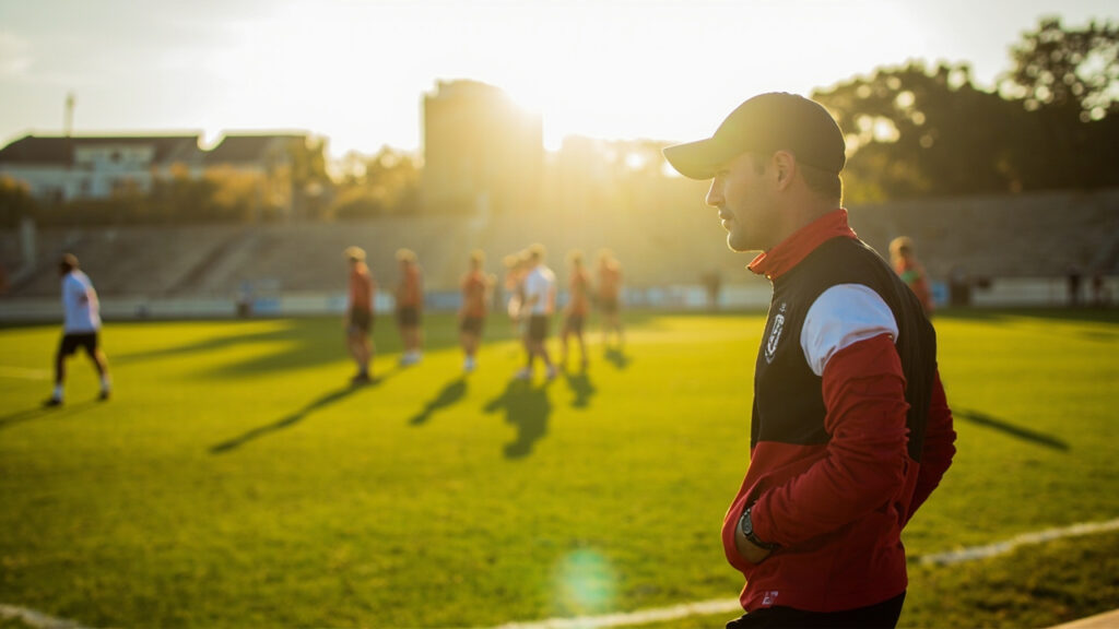 A football coach in training kit standing on the touchline of a football pitch during a training session.