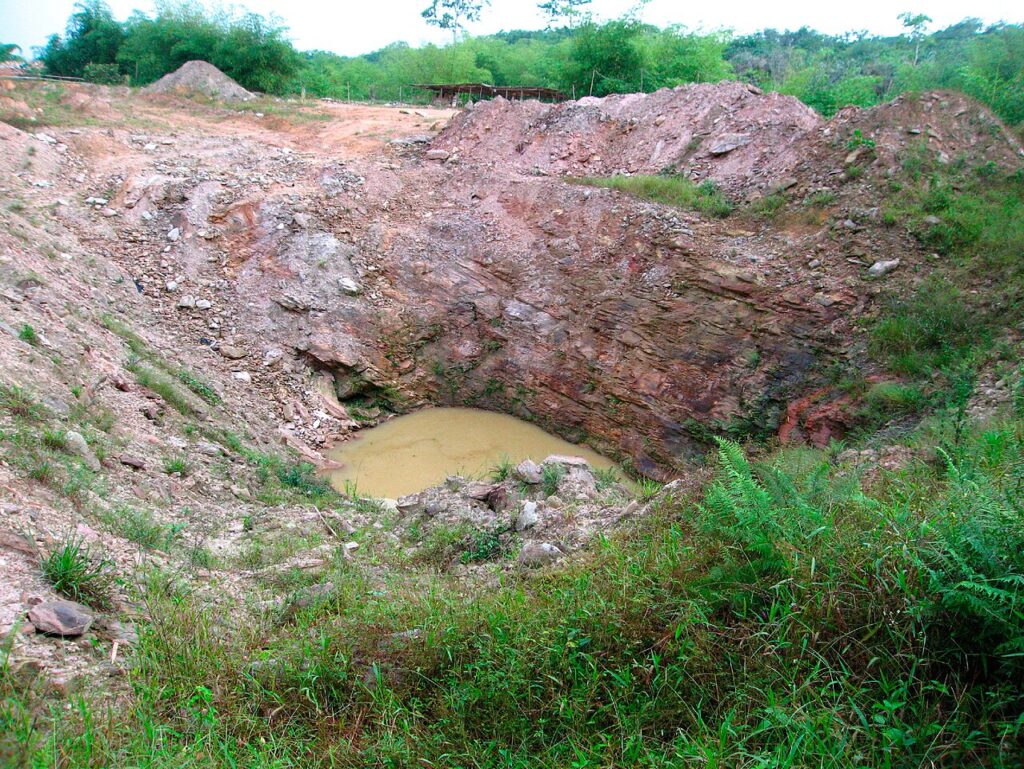An open-pit mining landscape with rust-coloured tailings heaps and processing equipment in a rural Ghanaian setting.