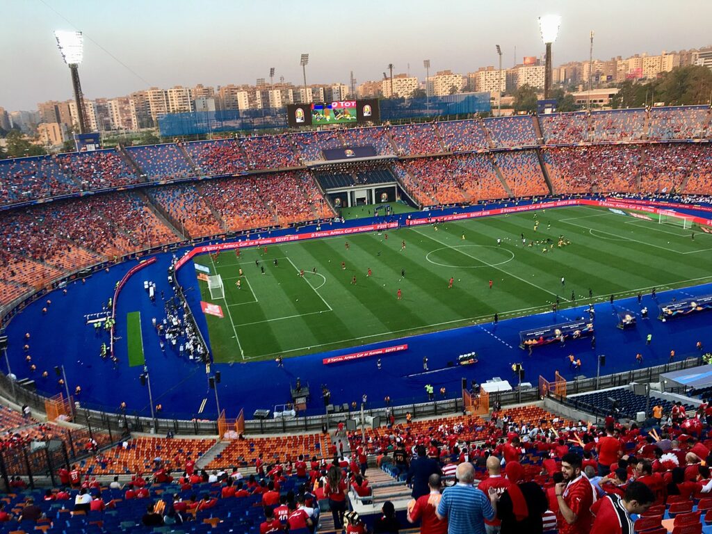 An empty outdoor football stadium at golden hour with green grass pitch, empty spectator seating, and floodlight towers.