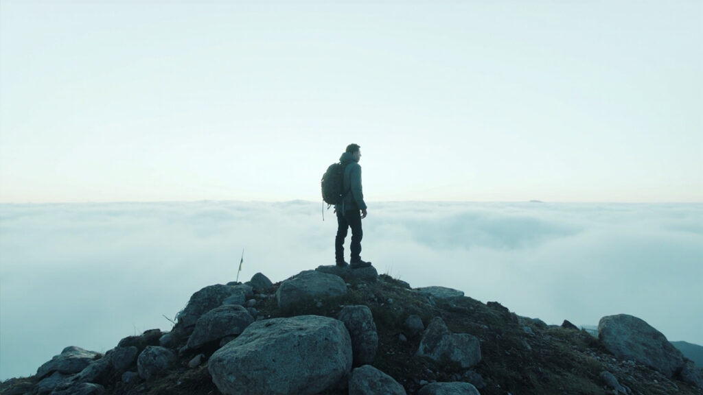 A lone hiker viewed from behind standing on a bare mountain summit above a sea of low cloud, with open landscape stretching ahead.