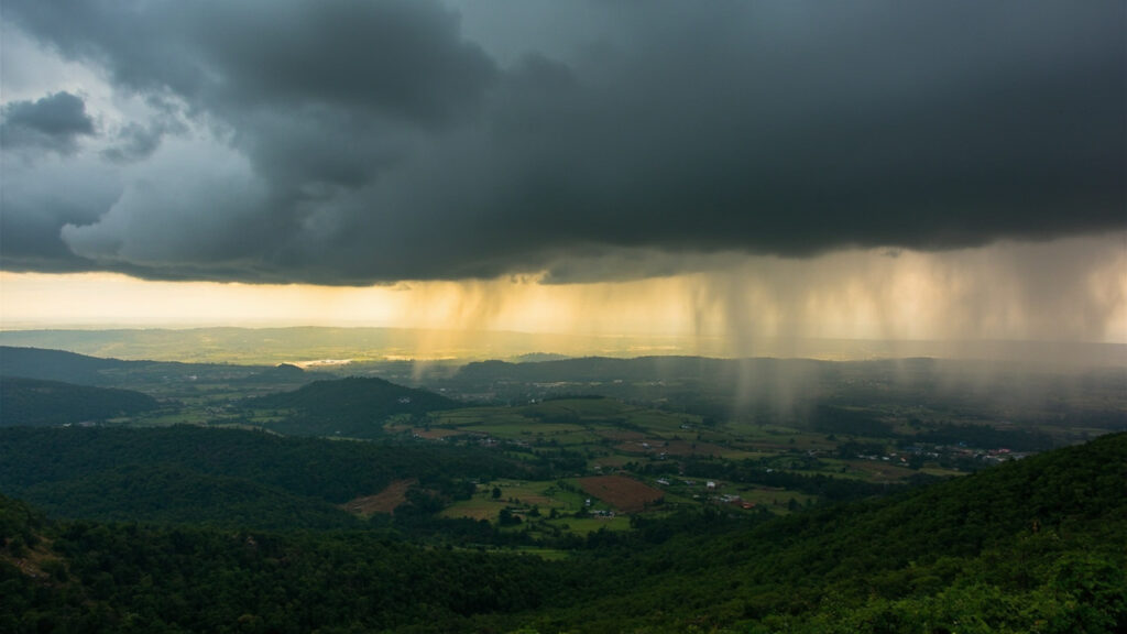 Heavy grey rain clouds gathering over rolling green hills in the Kenyan highlands, with visible rain shafts falling in the distance.