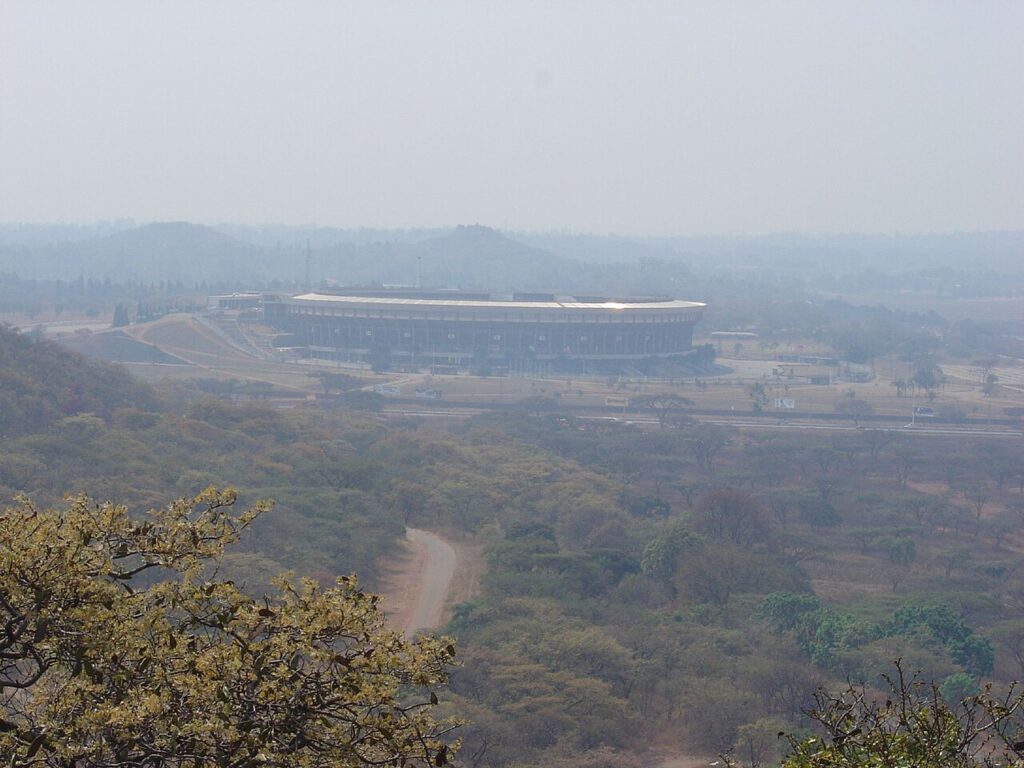 A wide daytime exterior view of the National Sports Stadium in Harare, showing the entrance, surrounding wall and tall floodlight towers.